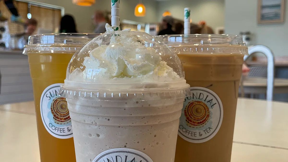 Three iced drinks in Sundial Coffee & Tea cups on a table inside the cafe, customers in the background.