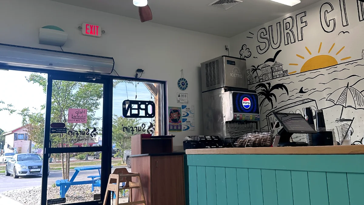 Interior of Surfin Hot Chicken with a Surf City beach mural, a soda fountain, and an OPEN sign on the door.