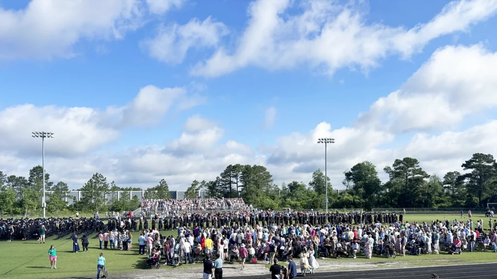 Graduates in black caps and gowns line the football field at Topsail High School while a large crowd of families fills the infield and packs the bleachers behind them.