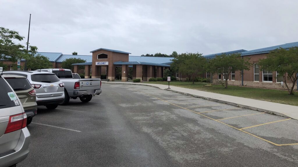 The front entrance of North Topsail Elementary School, a single-story brick building with a blue metal roof and a covered walkway, seen across the parking lot under an overcast sky.