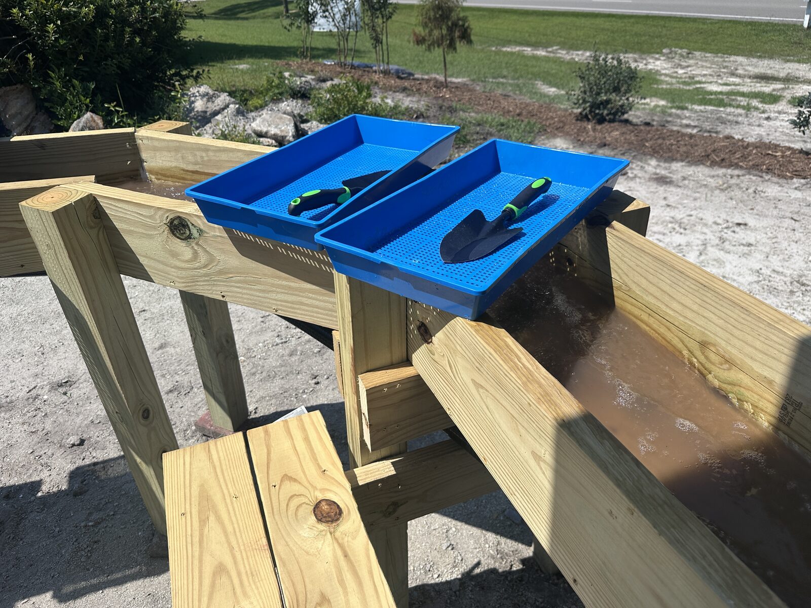 A wooden gem mining sluice with running water and blue plastic sifting screens holding hand trowels, set up outside on a sunny morning