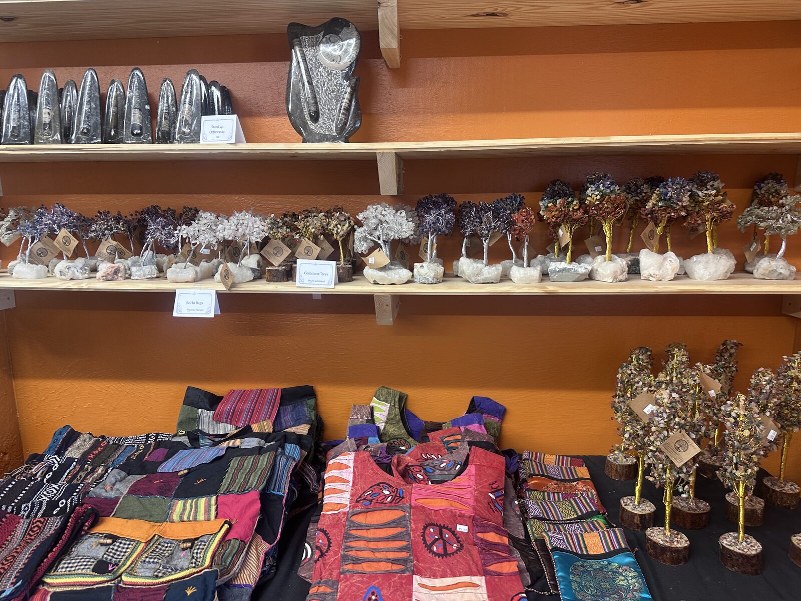 Wall shelves lined with wire gemstone trees and polished orthoceras fossils above a table of patchwork vests and bags