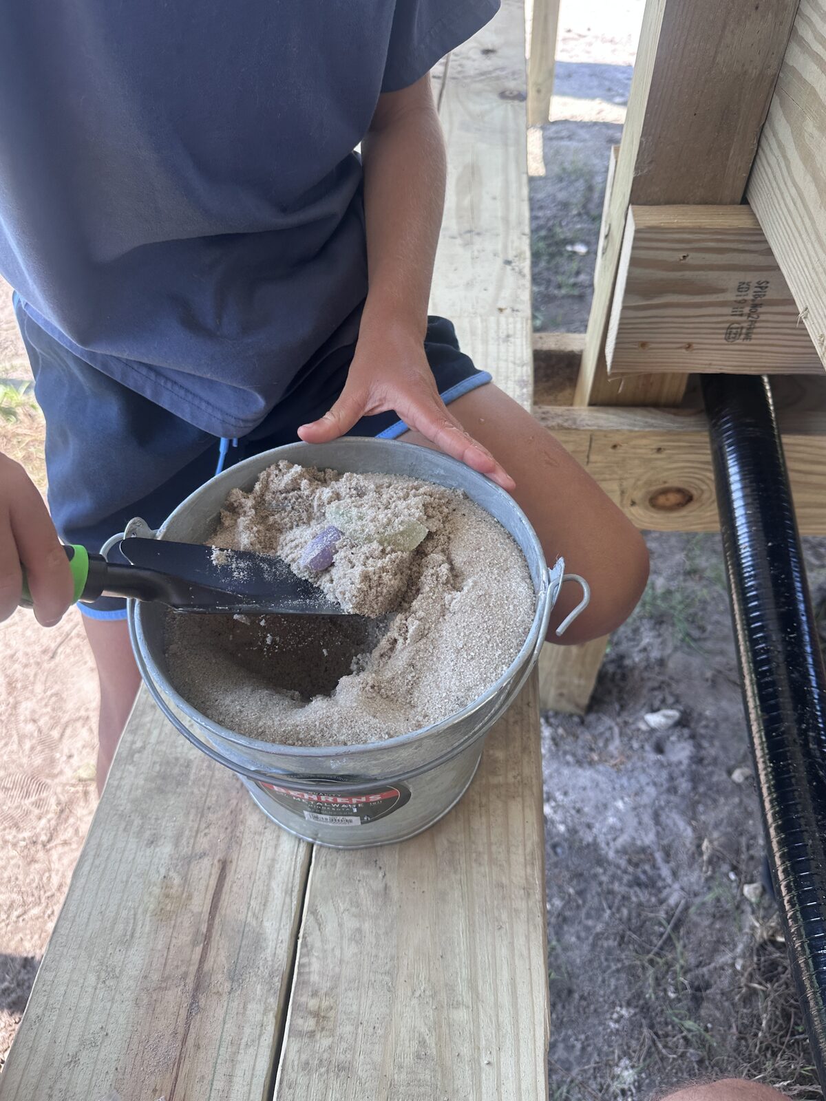A child in a blue shirt scooping sand from a galvanized bucket with a hand trowel, a green crystal already visible in the material